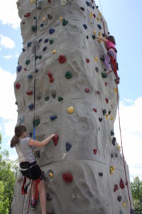 Two young girls are rock climbing on an outdoor climbing wall. The wall is tall and equipped with various colored holds. Both girls are secured with harnesses and climbing ropes. There is a blue sky and green trees in the background.