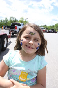 A smiling young girl with face paint and a sticker in a parking lot with trees and vehicles in the background. She has a lightning bolt on her forehead and paw prints on her cheeks. She wears a light-blue shirt with a colorful design.