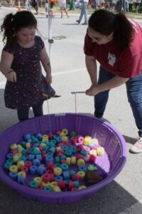 A young girl and an adult are playing a game involving fishing rods with strings, attempting to catch floating objects in a purple plastic pool filled with colorful foam pieces at an outdoor event. Other people and event activities are visible in the background.