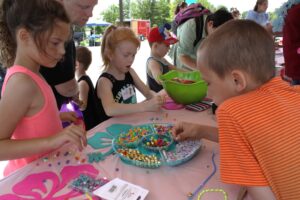 Children and adults participate in a bead crafting activity at an outdoor event. The table is covered with various colorful beads and crafting supplies. Some children are stringing beads while others look through the assortment to choose their favorites.