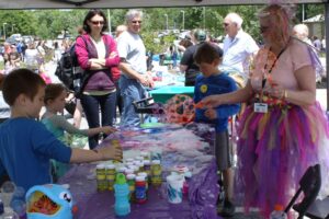 A group of children are gathered around a table filled with bubble-making supplies outdoors. An adult dressed in a colorful, fairy-like costume is helping the children create large bubbles. Several adults stand in the background, watching the activity.