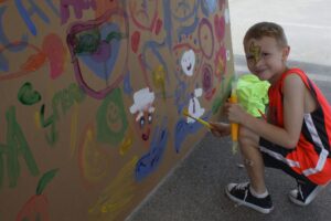 A young boy with face paint wearing an orange and black outfit is crouching and painting on a large piece of paper or canvas attached to a wall. The painting includes various colorful shapes and characters. The boy is holding a neon green shirt in one hand.