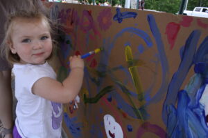 A young child stands in front of a large piece of cardboard covered with colorful paint markings. The child, holding a paintbrush in one hand, looks at the camera and smiles. Various abstract shapes and lines in different colors are visible on the cardboard.