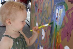 A young child with short blonde hair in a green floral dress holds a paintbrush and applies colorful paint to a large, upright canvas filled with various abstract shapes and colors. Their expression shows concentration and engagement in the painting activity.