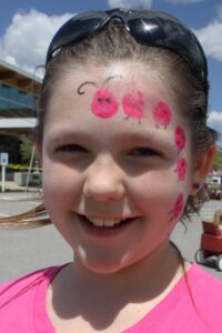 A young girl with a big smile has ladybug face paint on her forehead and cheek. She wears sunglasses pushed up on her head and is standing outdoors, with a building in the background and a red wagon behind her. The sky is blue with some clouds.