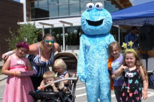 A group of children and an adult gather around someone in a blue, furry costume with large googly eyes, resembling the Cookie Monster character from Sesame Street. They are outdoors at a community event, with tents and balloons visible in the background.