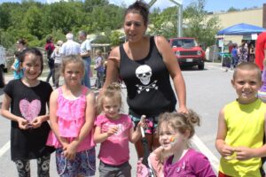 A woman stands with five children in a sunny outdoor setting. The woman, wearing a black tank top with a skull design, has her arms around some of the children. The children are smiling; one has a face paint rainbow. There are people, cars, and trees in the background.