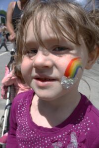 A young child with light brown hair and a purple shirt smiles at the camera. She has a colorful rainbow painted on her cheek and a small piece of plastic wrap under it. In the background, people are walking and pushing strollers. The sky is blue.