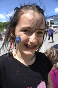 A young girl with windblown hair smiles at the camera. She has a blue creature face painting on her cheek. She is wearing a black T-shirt and is outdoors with a crowd visible in the background. A sunny sky and trees can be seen.