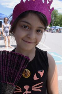 A young girl smiles at the camera, wearing a glittery purple crown and holding a black and purple fan. She has a giraffe pin on her black shirt, which features a pink cat face design. People and tents are blurred in the background, suggesting an outdoor event.