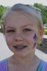 A young girl with blond hair smiles at the camera. She has a purple flower painted on her right cheek and is wearing a colorful top. The background includes greenery and a paved path, suggesting an outdoor setting.