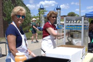 Two women wearing aprons and sunglasses are standing next to a popcorn machine at an outdoor event. One is operating the machine while the other smiles at the camera. A tree, brick building, and whimsical playground equipment are visible in the background.