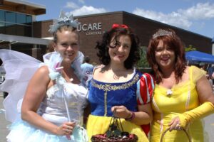 Three women are dressed as fairy tale characters; the first is a fairy in a white dress with wings and a wand, the second is in a Snow White costume with a basket, and the third is in a yellow gown with a tiara. They are outdoors in front of a building on a sunny day.