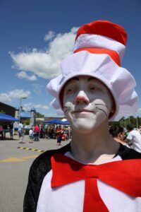 A person dressed as the Cat in the Hat, with face painted white and wearing a red and white striped hat and red bow tie, stands outdoors at a sunny event. A small crowd and blue tents are visible in the background under a partly cloudy sky.