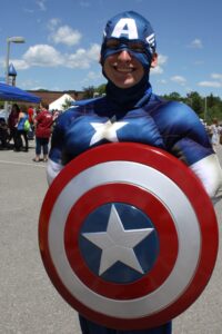 A person dressed in a Captain America superhero costume smiles while holding a red, white, and blue shield. The background shows a sunny outdoor event with a few people and tents visible.