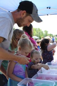 A man and two young girls engaged in an outdoor activity under a tent, with containers of water in front of them. The man assists one girl while the other reaches into a nearby container. Other children and adults are visible in the background.