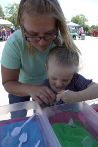 A woman and a young child participate in an outdoor sand art activity. The woman, wearing glasses and a light green shirt, helps the child, who is holding a small spoon and scooping colorful sand into a container. Tubs of green and blue sand are on the table.