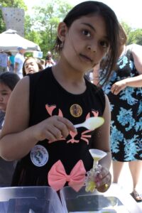 A young girl with long dark hair scoops sand into a small container at an outdoor event. She is wearing a black dress with pink accents and has a few stickers on her outfit. Other children and adults are visible in the background under a sunny sky.