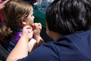 A young child sits patiently as an artist paints a yellow smiley face on her cheek at an outdoor event. The child is wearing a purple outfit, while the artist concentrates on the face painting. In the background, a table with water bottle and papers is visible.