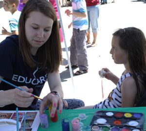 A woman is face painting a young girl at an outdoor event. The girl is holding an ice cream cone while the woman uses a brush to apply paint. Various paint supplies are on the table in front of them. Other people are visible in the background.