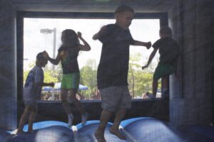 Four children are jumping inside a blue inflatable bounce house. They are silhouetted against the light coming through the mesh walls. The children appear to be having fun as they bounce and interact with each other. Trees and a crowd can be seen in the background.