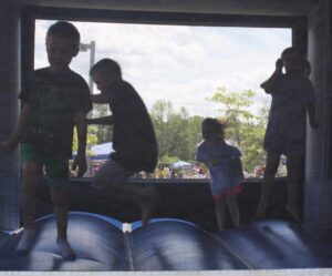 Children jump on an inflatable bounce house at an outdoor event. The sunlight creates a backlit effect, casting their figures in shadow. Trees and a crowd of people can be seen in the background.