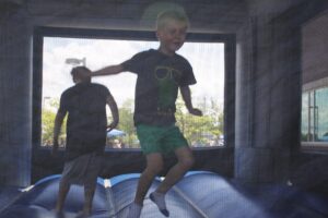 A young boy with blond hair and a black shirt featuring a green graphic jumps on a blue inflatable bounce house. An adult in a gray shirt is also in the bounce house. The scene is seen through a mesh safety net, with a sunny outdoor backdrop.
