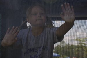 A child with light-colored hair in pigtails presses their hands against a mesh screen. They are wearing a gray shirt with blue text that appears partially obscured. The background shows an outdoor setting with trees and blue sky visible.
