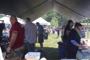 People working together under a large tent at an outdoor event. Some individuals are preparing food on grills and tables, while others are serving or organizing. The surrounding area features more tents and attendees enjoying the sunny day.
