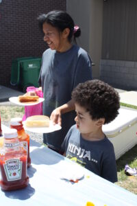 A woman and a young boy stand at an outdoor table with plates of hot dogs. The woman smiles, and the boy looks down at the table. Bottles of ketchup and mustard are on the table. They appear to be at a casual gathering or picnic with a building and cooler in the background.