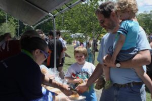 A man holding a small child and a young boy stand at a food stall outdoors. The man is receiving food from a vendor under a canopy. Other people are visible in the background, suggesting a community event or a festival in a park setting.