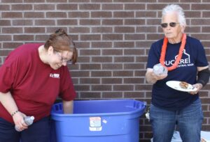 Two people are next to a large blue bin in front of a brick wall. The person on the left, wearing a red shirt, is bending over and reaching into the bin. The person on the right, wearing sunglasses and a dark shirt with an orange lei, holds a water bottle and a plate of food.