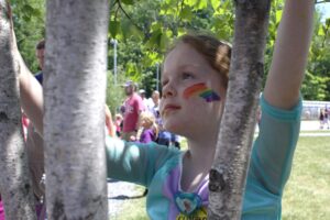 A young girl with a rainbow painted on her cheek reaches up to touch the branches of a tree. She is wearing a light blue top and appears to be in a park with people and greenery in the background. The weather is sunny.