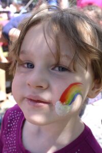 A young child with brown hair and blue eyes smiles at the camera. There is a colorful rainbow with a cloud painted on their cheek. They are wearing a purple shirt with white polka dots. The background shows a blurred outdoor setting with people.