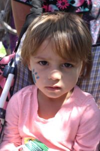 A young child with light brown hair and a serious expression sits in a plaid stroller. The child wears a pink shirt with blue face paint in the shape of a flower petal on their right cheek, peering directly at the camera.