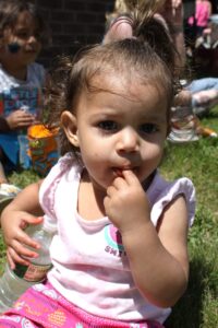 A toddler with brown hair pulled into a small ponytail on top of their head sits on the grass, holding a bottle of water. The child is wearing a light pink shirt and colorful shorts, and is looking directly at the camera while putting fingers in their mouth. Other children are visible in the background.