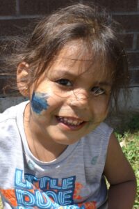 A young child with long dark hair smiles at the camera. Blue and green paint smudges are on their cheek, with a few streaks on their forehead and chin. The child is wearing a light gray tank top printed with "LITTLE DUDE" in colorful letters.