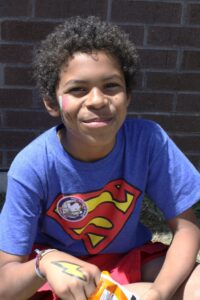 A child with curly hair sits outside against a brick wall, wearing a blue T-shirt with a red and yellow Superman logo. They have face paint of a pink heart and a yellow lightning bolt on their arm. The child is smiling and holding a bag of snacks.