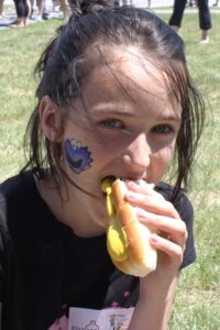 A young girl is enjoying a hot dog with mustard at an outdoor event. She has a painted blue character on her cheek and is wearing a black t-shirt. Other people and activities are visible in the blurred background on a grass field.
