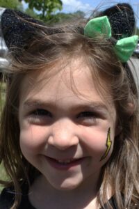 A young girl with brown hair smiles at the camera. She wears a pair of black cat ears with a green bow and has a painted yellow lightning bolt on her cheek. The background is an outdoor setting with trees and grass.