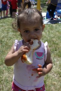 A young child with dark hair in a ponytail is standing outdoors, holding and eating a hot dog in a bun. The child is wearing a pink tank top with a watermelon graphic and "SMILE" text, and has some ketchup stains on their shirt. Other people are visible in the background.