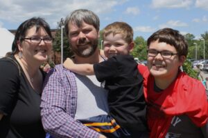 A smiling family poses outdoors on a sunny day. From left to right, a woman wearing glasses, a man with gray hair and a beard, a young child in the man's arms, and an older child with glasses and a red jacket. Trees and a building are visible in the background.