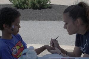 A young child with face paint wearing a Superman shirt sits at a table outdoors. An adult sits across from them, carefully painting the child’s hand. There are art supplies and a water bottle on the table, with greenery in the background.