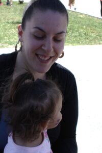 A woman with a ponytail and large hoop earrings smiles down at a young child with curly hair and a small purple mark on their cheek. They are outdoors, with grass and a lighter paved area in the background.