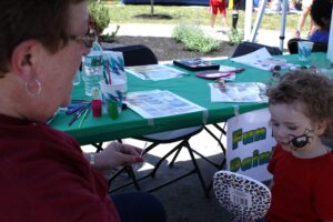 A child with face paint sits at a table holding a decorated paper plate, while an adult with short hair and glasses interacts with them. The table is covered with a green tablecloth and assorted craft supplies. In the background, people are gathered outdoors.