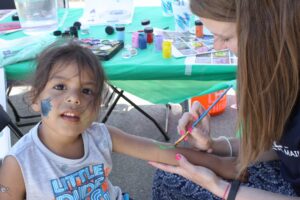 A young child with face paint sits at a table outdoors, watching attentively as an adult paints their arm. The table is covered with a green tablecloth and various paint supplies. The child wears a sleeveless shirt, and the adult is focused on the painting task.