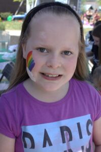 A young girl with long hair smiles at the camera. She has a painted rainbow on her cheek and is wearing a purple shirt with a partially visible graphic design. The background shows an outdoor setting with trees, tables, and other people.