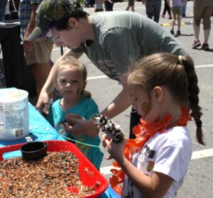 A young man assists two small children at a table covered in birdseed. One girl, wearing an orange lei and face paint, examines a pine cone. The second girl watches intently while holding something. They are participating in an outdoor craft activity.