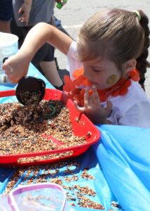 A young girl plays with birdseed at an outdoor event. She uses a scoop to transfer the seeds into a tray. She wears an orange flower necklace and has a smiley face sticker on her cheek. There are other people in the background and a blue cloth covers the table.