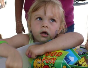 A young child with blond hair leans on a table, holding colorful brochures that read "Food Fun." The child has a slightly smudged face and looks to the side with a curious expression. An adult wearing a pink shirt stands in the background.